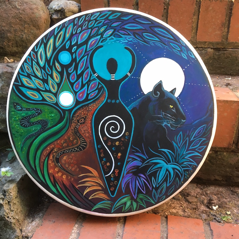 May include: A circular, hand-painted artwork depicting a central female form with raised arms, a tree, and a panther. The piece uses blues, greens, and browns, with a white border. The design incorporates symbolic imagery, with a full moon in the background.