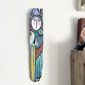 Könnte beinhalten: Ein handbemaltes Holzkunstwerk, das eine stilisierte Figur darstellt. Das Kunstwerk zeigt ein Gesicht mit rosa Wangen, einer blauen Krone und komplizierten Mustern in Rot, Blau und Gelb. Das Kunstwerk ist an einer weißen Wand befestigt.