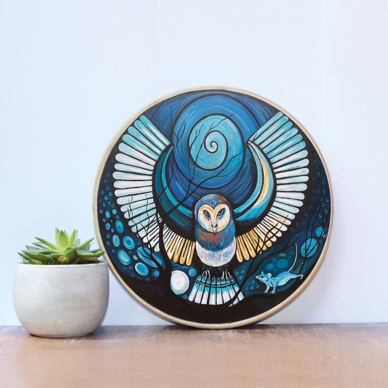 May include: Round wall art featuring a stylized owl with outstretched wings, painted in shades of blue, white, and gold. The owl is centered against a dark background with a spiral design. A small succulent plant in a white pot is to the left.