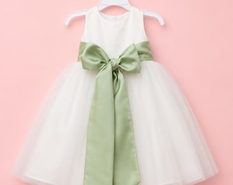 Puede incluir: Un vestido blanco sin mangas con cuello redondo y una falda de tul con capas. Un fajín ancho de satén verde salvia está atado en un lazo grande y prominente en la cintura, con largas cintas que caen por la parte delantera de la falda.