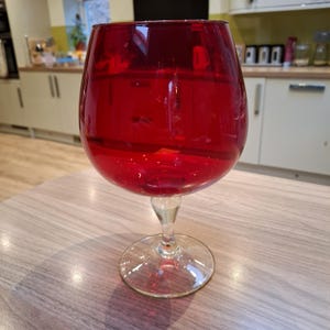 May include: A large, round, deep red glass goblet with a wide, bulbous bowl and a clear, transparent stem and circular base. The glass features a smooth, polished surface that reflects light, showcasing a vibrant, saturated crimson colour throughout.