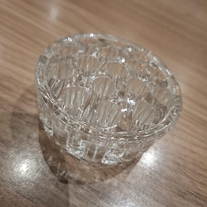 May include: A clear, circular glass flower frog featuring a series of vertical cylindrical openings arranged in a grid pattern. This heavy, transparent glass accessory is designed with a flat base and a textured, multi-holed top surface.