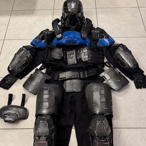 Puede incluir: Un conjunto completo de armadura futurista negra y gris, que incluye un casco, peto con detalles azules, hombreras, protectores de brazos, armadura de piernas y guantes. La armadura se muestra sobre una superficie embaldosada, destacando su diseño detallado.