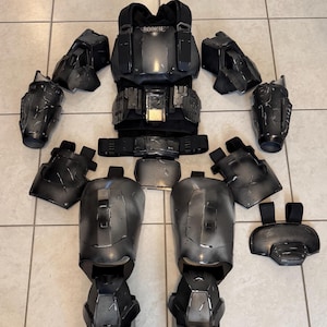 May include: A full set of black and grey armour components, including a chest plate with the text "ROOKIE", shoulder pads, arm guards, leg armour, and a belt. The armour appears to be made of a hard, durable material.