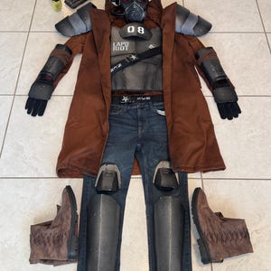 May include: A complete cosplay costume laid out on a tiled floor. The costume includes a brown duster coat, a black helmet with a red visor, a chest plate with "08" and "LAPD RIOT" text, blue jeans, and brown boots. Additional accessories are also present.