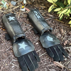 K&ouml;nnte beinhalten: Paar schwarze und graue Armsch&uuml;tzer mit Handschuhen, mit einem Stern und einem Faust-Emblem. Auf den Armsch&uuml;tzern steht das Wort "ZOR" als Schablone. Die Handschuhe sind schwarz und scheinen aus strapazierf&auml;higem Material zu sein. Auf einer Unterlage aus Holzsp&auml;nen.