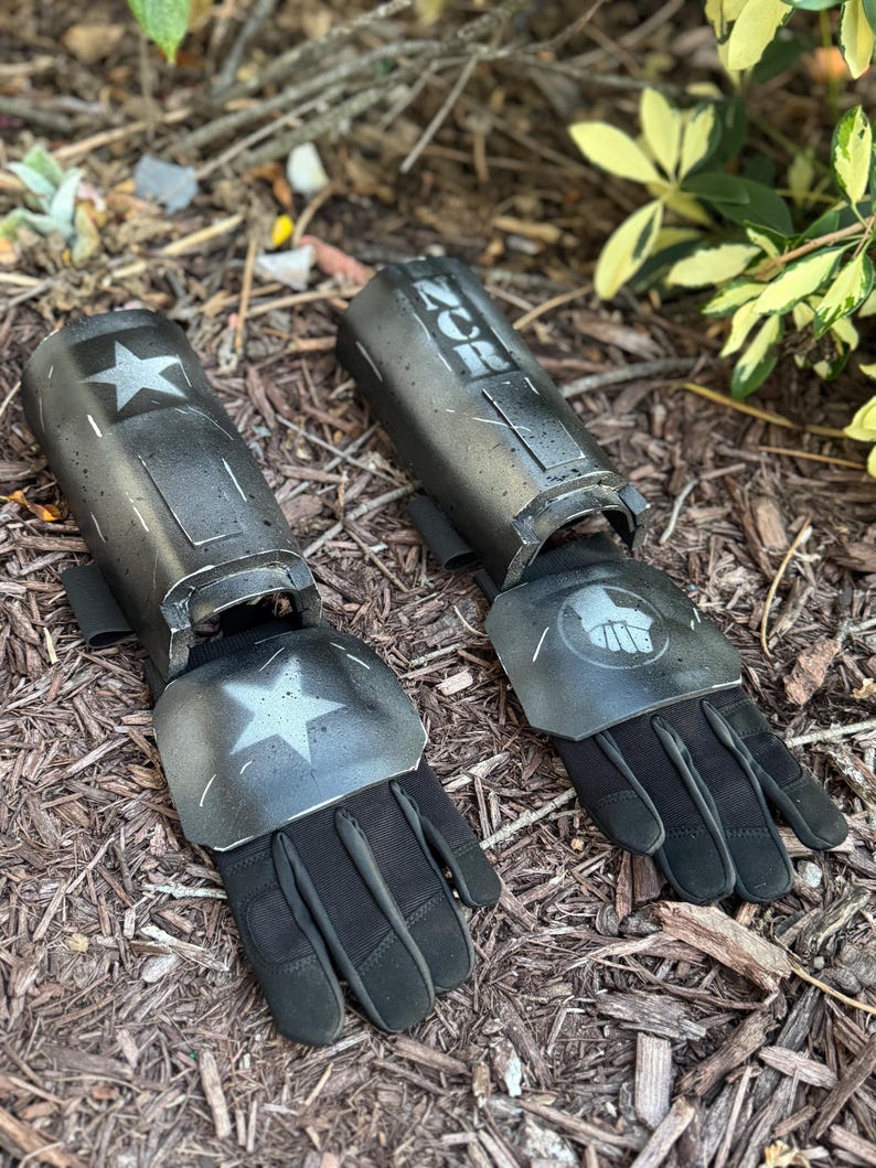 K&ouml;nnte beinhalten: Paar schwarze und graue Armsch&uuml;tzer mit Handschuhen, mit einem Stern und einem Faust-Emblem. Auf den Armsch&uuml;tzern steht das Wort "ZOR" als Schablone. Die Handschuhe sind schwarz und scheinen aus strapazierf&auml;higem Material zu sein. Auf einer Unterlage aus Holzsp&auml;nen.