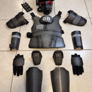 K&ouml;nnte beinhalten: Ein komplettes Set schwarzer Aufruhr-Ausr&uuml;stung, einschlie&szlig;lich eines Helms mit rot get&ouml;ntem Visier, einer Weste mit dem Aufdruck "LAPD RIOT" und "08", Arm- und Beinschutz, Handschuhen und anderem Zubeh&ouml;r. Die Ausr&uuml;stung ist auf einer hellen Oberfl&auml;che angeordnet.