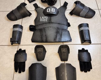 Könnte beinhalten: Ein komplettes Set schwarzer Aufruhr-Ausrüstung, einschließlich eines Helms mit rot getöntem Visier, einer Weste mit dem Aufdruck "LAPD RIOT" und "08", Arm- und Beinschutz, Handschuhen und anderem Zubehör. Die Ausrüstung ist auf einer hellen Oberfläche angeordnet.
