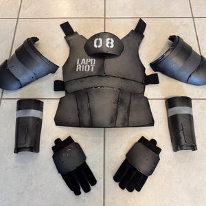 K&ouml;nnte beinhalten: Ein komplettes Set grauer Riot-Ausr&uuml;stung, einschlie&szlig;lich eines Brustschutzes mit "LAPD RIOT" und der Nummer "08", zwei Armsch&uuml;tzern, zwei Beinsch&uuml;tzern und zwei Handschuhen. Die Ausr&uuml;stung ist auf einer gefliesten Oberfl&auml;che angeordnet.
