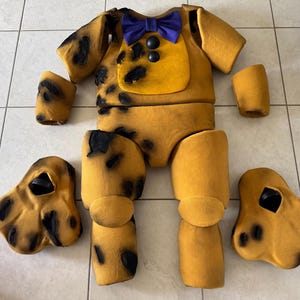May include: A disassembled, tan-colored costume with black spots, a yellow chest, and a purple bow tie. The costume includes a torso, arm sleeves, leg pieces, and two shoe-like pieces. The costume is likely for a character or mascot.