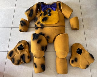 May include: A disassembled, tan-colored costume with black spots, a yellow chest, and a purple bow tie. The costume includes a torso, arm sleeves, leg pieces, and two shoe-like pieces. The costume is likely for a character or mascot.