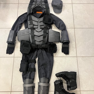 Op de afbeelding: Een zwart tactisch pak met een grijs gepantserd vest, zwarte handschoenen, zwarte laarzen en een grijze pet. Het pak heeft een aantal zakken en banden.