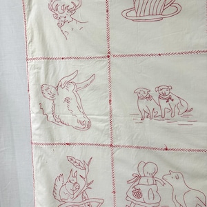 May include: A white fabric quilt featuring a grid of red embroidered illustrations including a deer, a teacup, a cow, two dogs, a squirrel, and a person with a lamb. The sections are joined by red cross-stitch embroidery along the seams.