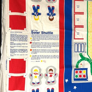 May include: A fabric panel featuring a colorful space-themed design. It includes illustrations of astronauts, bunk beds, and text that reads "Space Race Solar Shuttle." The panel also has sewing instructions and red pocket cutouts.