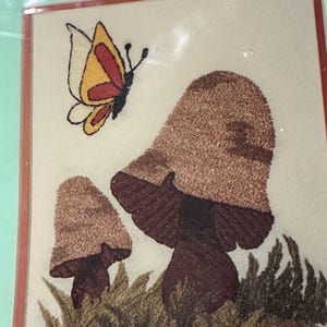 May include: A square wall art piece featuring a textured embroidery design of two brown mushrooms growing in green grass with a yellow, white, and red butterfly flying above them, all enclosed within a thin red border on a light background.