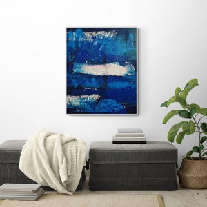 May include: Abstract painting with blue and white brushstrokes on a canvas. The painting is framed and hanging on a white wall above a gray ottoman.