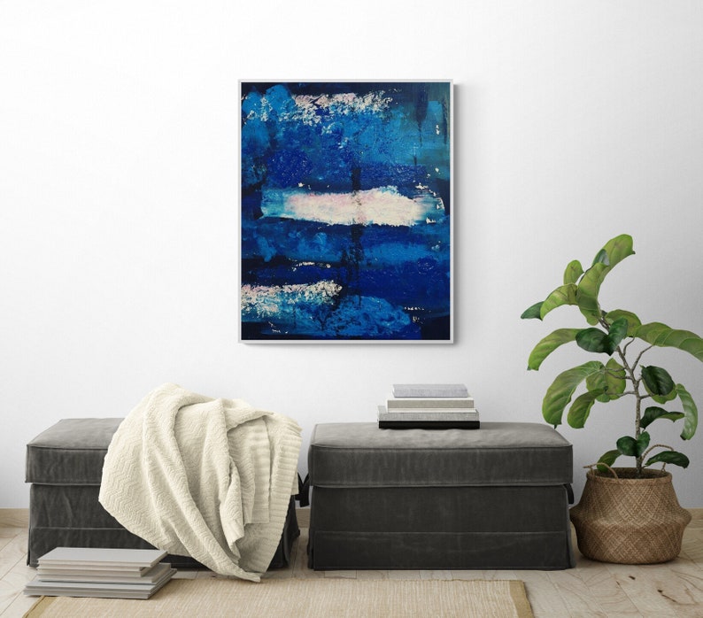 May include: Abstract painting with blue and white brushstrokes on a canvas. The painting is framed and hanging on a white wall above a gray ottoman.