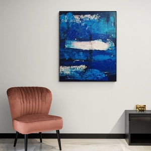 May include: A pink velvet armchair with a black frame sits in front of a white wall with a framed abstract painting of blue and white brushstrokes.