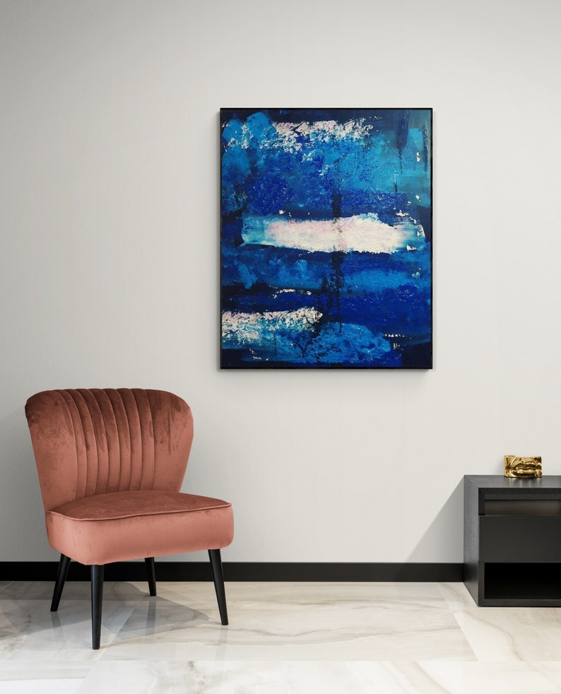May include: A pink velvet armchair with a black frame sits in front of a white wall with a framed abstract painting of blue and white brushstrokes.