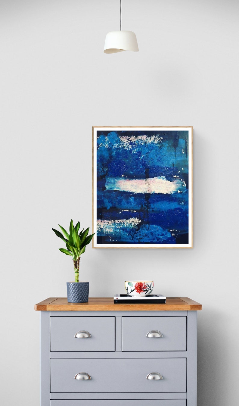 May include: A framed abstract painting with blue, white, and pink brushstrokes. The painting is hanging on a white wall above a gray dresser with four drawers.
