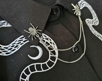 Könnte beinhalten: Ein silberner Spinnenkragen-Pin mit Ketten-Detail, befestigt an einem schwarzen Hemd. Das Hemd hat weiße, gestickte Schlangen- und Mond-Designs. Die Spinnen-Pins befinden sich am Kragen.