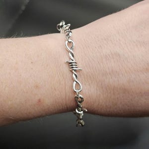 May include: A silver-coloured bracelet featuring a continuous design of twisted metal links that mimic the appearance of barbed wire. The metallic chain consists of repeating segments with sharp, protruding spikes, creating a textured and industrial aesthetic.