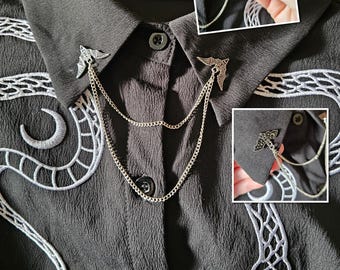 May include: A black button-down shirt with white embroidered snake and moon designs. The shirt collar features silver-toned wing-shaped brooches connected by delicate chains. The shirt has a textured appearance.