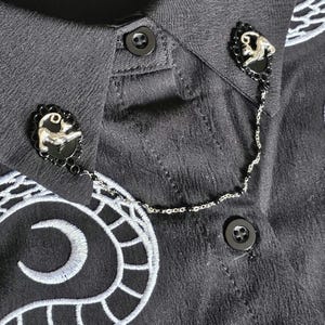 May include: Close-up of a black shirt featuring decorative silver-toned monkey-shaped brooches linked by a delicate chain. The brooches have black oval frames with a scalloped edge. The shirt has black buttons and white embroidered details.