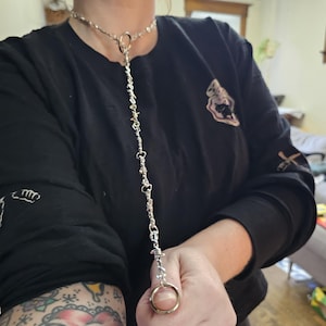 Op de afbeelding: Zilverkleurige ketting met een choker-ontwerp, met een centrale ring en een ketting die uitloopt in een vingerring. De ketting wordt gedragen over een zwart shirt met lange mouwen. Het ontwerp bevat kleine, decoratieve elementen langs de ketting.