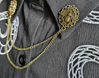May include: A vintage-style bronze bee brooch with a decorative frame and chain detail. The bee is centered in an oval frame. The brooch is attached to a black shirt with white embroidery.