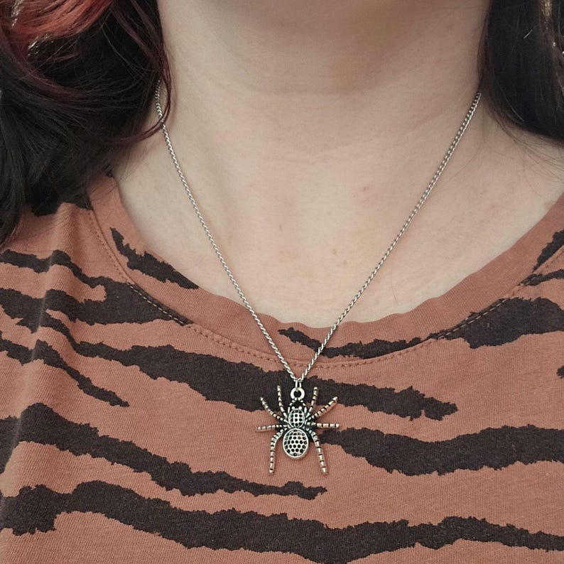 May include: Silver-toned spider pendant necklace. The spider charm has detailed legs and a textured body. The necklace is worn on a person with a brown and black patterned shirt.
