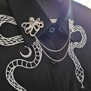 May include: A black shirt featuring white embroidered snake and crescent moon patterns. Two silver brooches, an octopus and a figure, are attached to the collar with fine chains. The shirt has a button-down front.