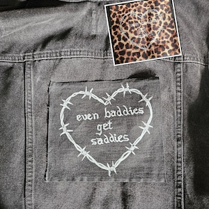 Pode incluir: Casaco de ganga cinzento escuro com um remendo com um coração de arame farpado e o texto "even baddies get saddies". Um remendo menor com um desenho de coração com estampa de leopardo também é visível.