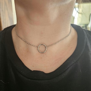 Puede incluir: Collar gargantilla plateado con una cadena fina y un colgante circular. El collar se lleva sobre una parte superior negra, creando un contraste que resalta el diseño de la joya. Un accesorio sencillo y elegante.