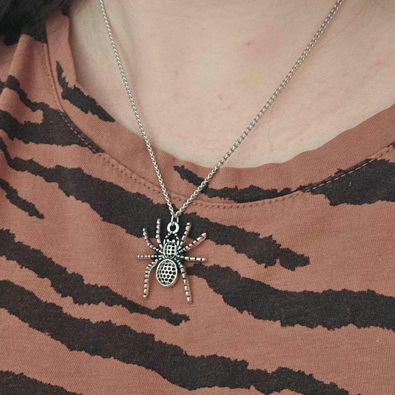 May include: Silver-toned spider pendant necklace. The pendant features a detailed spider design with eight legs and a textured body. The necklace is worn against a brown and black striped shirt.