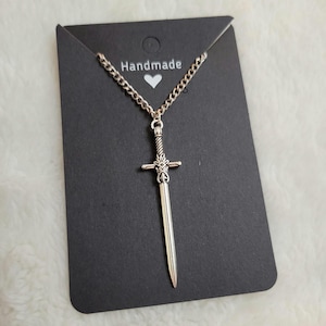 May include: A silver sword pendant necklace on a silver chain. The pendant is detailed with a cross at the top and a pointed tip. The necklace is displayed on a black card with the word "Handmade" written on it.