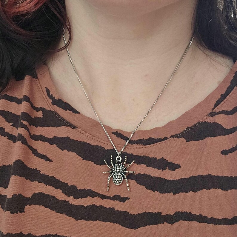 May include: Silver-toned spider pendant necklace. The spider charm has detailed legs and body, suspended from a delicate chain. The necklace is worn against a brown and black striped shirt.