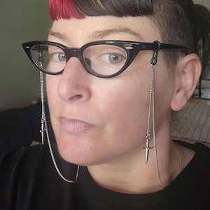 May include: Black cat-eye glasses featuring a silver-toned metal chain attached to each temple. The chain hangs down on both sides and terminates in a small, silver-coloured metal dagger charm. The glasses have clear lenses and a glossy finish.