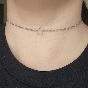 May include: A silver choker necklace with a star-shaped pendant. The necklace is made of a fine chain. The star pendant is positioned centrally on the chain, worn around the neck.