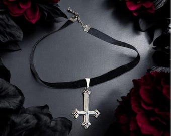 May include: A black velvet choker necklace with a silver inverted cross pendant. The cross has red accents and is suspended from a silver bail. The background is dark with red and black floral accents.