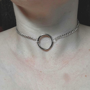 May include: A silver choker necklace with a large circular pendant. The necklace is made of a chain with a large ring in the centre. The necklace is worn around the neck.