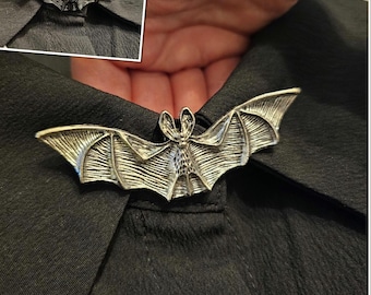May include: Silver-toned bat-shaped collar clip. The bat has detailed wing and body features, and is clipped onto a black collared shirt. The image includes a smaller inset view of the clip.
