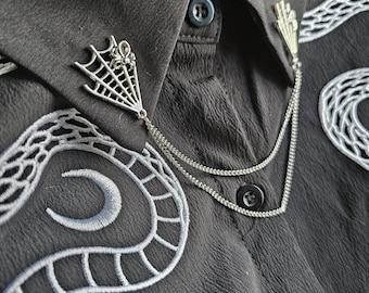 May include: Close-up of a dark gray shirt with silver spiderweb and spider collar pins. The shirt has white embroidered snake-like designs. The collar pins are connected by a delicate chain. The shirt has a button-down closure.