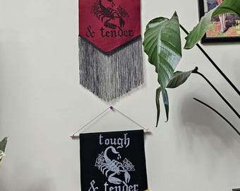 May include: Two wall banners hanging vertically. The top banner is deep red with black text reading tough & tender, a black scorpion, and black fringe. The bottom banner is black with white text reading tough & tender, a white scorpion, and gold fringe.