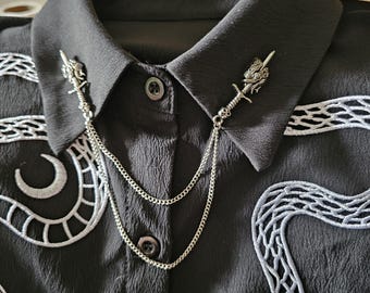 May include: A black shirt with white embroidered snake and moon patterns. Two silver-coloured collar pins, each with a sword and dragon design, are linked by a chain.
