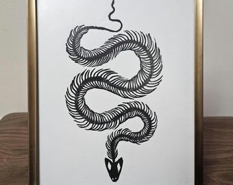 May include: A framed print featuring a black and white illustration of a snake skeleton. The snake is coiled, with its spine and skull clearly depicted. The frame is gold-colored, adding a touch of elegance to the artwork. The print is displayed on a wooden surface.