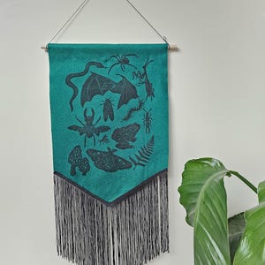 May include: Teal wall hanging with black fringe and a wooden dowel. The fabric features a repeating pattern of insects, bats, and other creatures. The wall hanging is suspended by a string from a small hook.