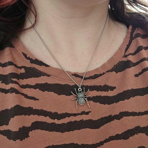 May include: A silver-colored spider pendant necklace. The spider has a detailed body and legs, suspended from a delicate chain. The necklace is worn against a brown and black striped shirt.