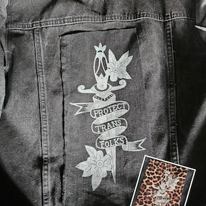 May include: Dark grey denim jacket featuring a white graphic on the back that reads "PROTECT TRANS FOLKS" with a dagger and floral design. A smaller leopard print patch with the same design is also visible.